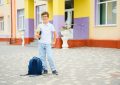 Un jeune écolier à lunettes pose fièrement devant son école avec son cartable à roulettes bleu au sol