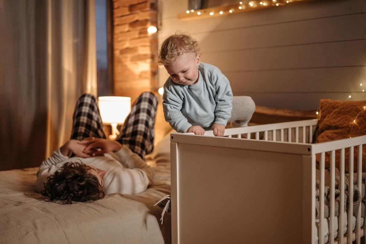Un enfant en bas âge se tient debout dans un lit parapluie blanc, un adulte allongé près de lui dans une chambre