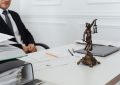 Homme en costume assis à un bureau avec une statue de la justice, des papiers et un ordinateur portable