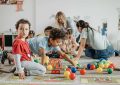 Enfants et adultes jouent avec des balles et jouets sur un tapis coloré dans une pièce blanche