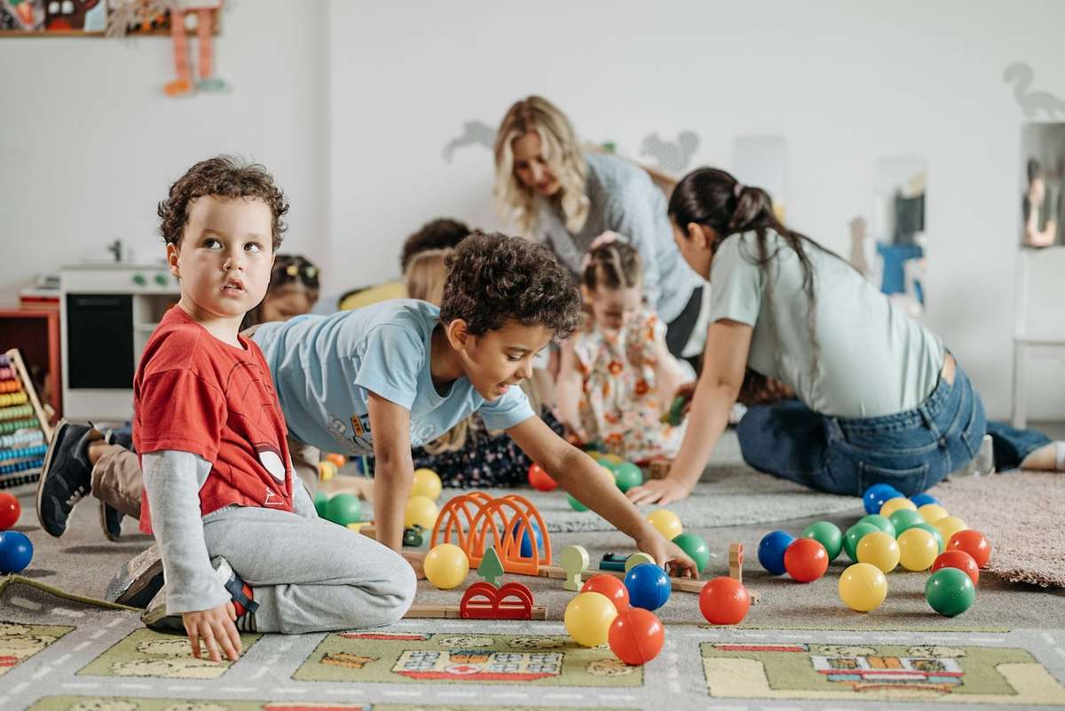 Enfants et adultes jouent avec des balles et jouets sur un tapis coloré dans une pièce blanche