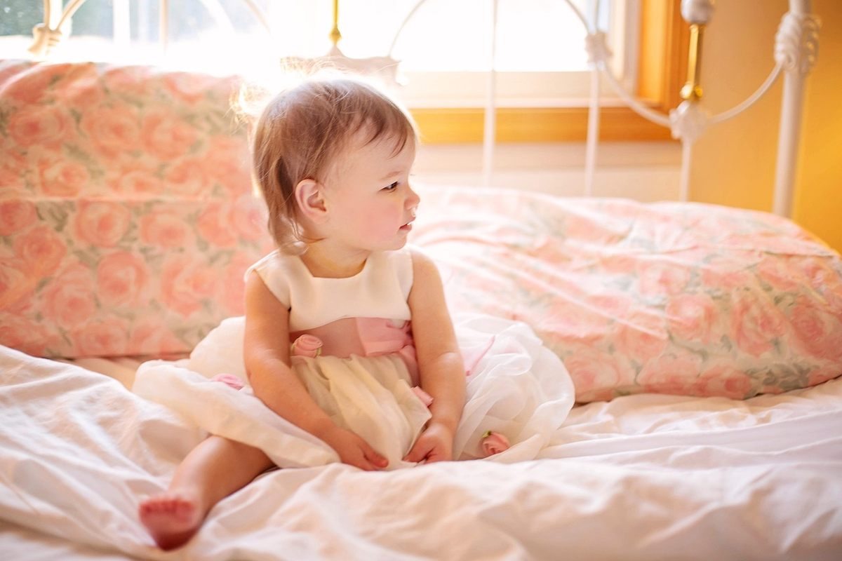 Petite fille en robe blanche assise sur un lit avec oreiller floral